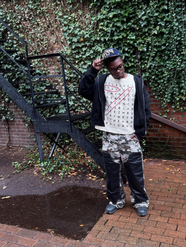 Customer in a black jacket over a printed tee and camo pants by an ivy wall