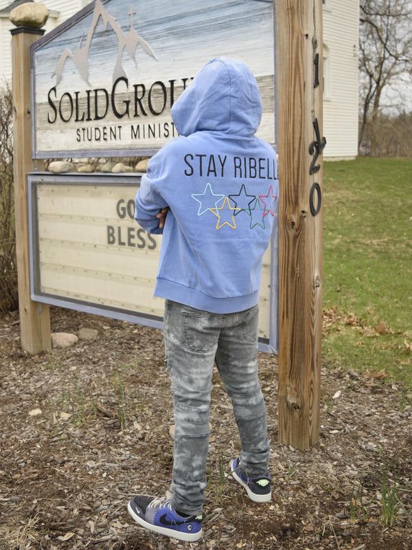 Back view of a periwinkle Stay Ribelle star hoodie beside a church sign